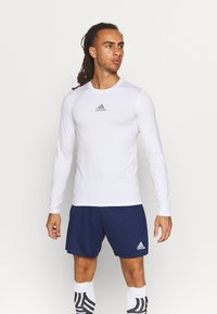 Weißes langärmliges Sporthemd mit einem grauen adidas-Logo, kombiniert mit navyblauen Shorts. Der Stoff wirkt glatt und feuchtigkeitsableitend.