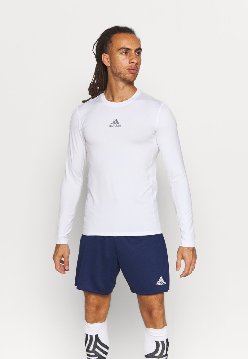 Weißes langärmliges Sporthemd mit einem grauen adidas-Logo, kombiniert mit navyblauen Shorts. Der Stoff wirkt glatt und feuchtigkeitsableitend.