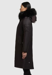 Lang sort parka jakke med en foring af pels i hætten, to sidetasker og en lommepose på ærmet; glat tekstur og elegant design.