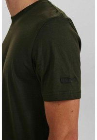 T-shirt a maniche corte verde oliva in tessuto morbido con scollo rotondo, caratterizzato da una toppa nera con logo sulla manica sinistra.