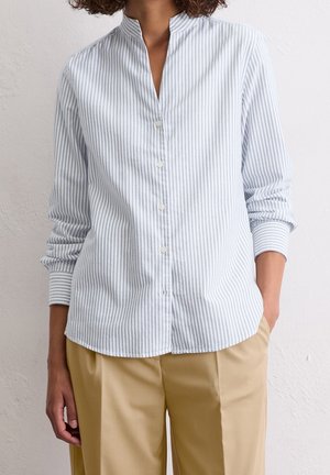 Vrouw draagt een lichtblauw-wit gestreept overhemd met verticale strepen en beige broek, staand tegen een effen muur met één hand in de zak.