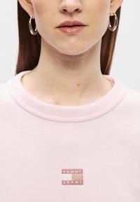 Różowa bluza z ribbowanym dekoltem, z małym haftowanym logo "TOMMY JEANS" na prostokątnym naszywce. Gładka faktura, casualowy styl.