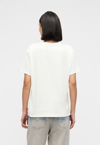 Γυναίκα με ίσια σκούρα μαλλιά που φοράει απλό λευκό κοντομάνικο μπλουζάκι και ανοιχτόχρωμο μπλε τζιν, φαινομένη από πίσω σε λευκό φόντο.