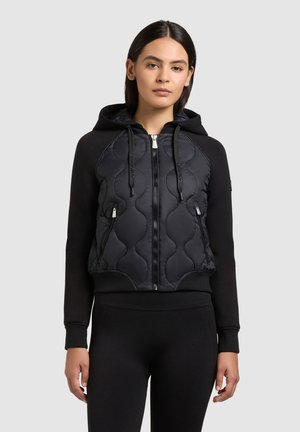 Kobieta w czarnej pikowanej bluzie z zamkiem, kieszeniami z przodu i ściągaczami, w połączeniu z czarnymi legginsami, stojąca na jasnym tle.