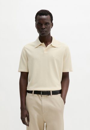Mann trägt beige kurzärmeliges Poloshirt, beige Hose und schwarzen Gürtel, steht mit einer Hand in der Tasche vor weißem Hintergrund.