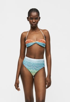 Femeie cu părul împletit, care poartă un top de bikini texturat, fără bretele, cu bretele halter, și chiloți de bikini cu talie înaltă, în tonuri gradient de albastru și verde.