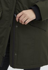 Veste isolée vert foncé avec fermeture éclair, boutons-pression et détails de poignets côtelés. Dispose d'une texture de tissu extérieur lisse.