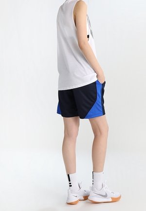Person steht mit einer Hand in der Tasche, trägt ein weißes ärmelloses Shirt, blau-schwarze Shorts, weiße Socken und weiße Sportschuhe.