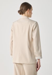 Beige kohandatud bleiser, mis on valmistatud siledast kangast. Omab struktureeritud siluetti, pikki varrukaid ja triibulisi mansette. Tagaküljelt on näha puhtaid jooni.