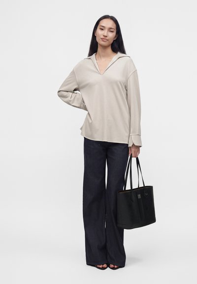 Jeune femme aux longs cheveux noirs porte un top beige ample et un pantalon noir large, tenant un sac cabas noir, debout devant un fond clair uni.