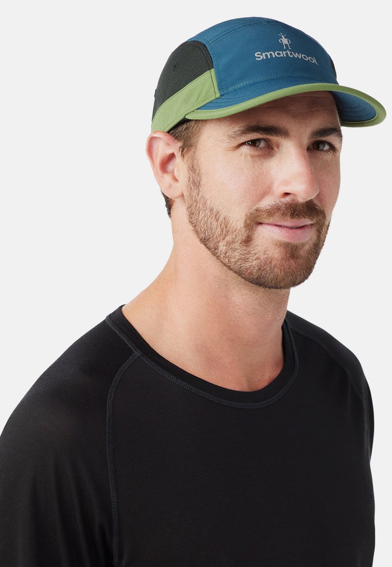 Baseball cap in blue with green accents and black mesh panels. Logo on front. Worn with a black shirt, showing a curved bill design.