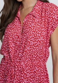 Rotes Kleid mit Kragen und kurzen Ärmeln, das ein pinkes Tiermuster aufweist und an der Taille mit einem passenden Gürtel gebunden ist. Weicher Stofftextur.