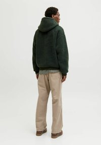 Haut en molleton vert avec un tissu texturé, coupe décontractée, ourlet et poignets côtelés, porté sur une chemise à carreaux, associé à un pantalon beige et des chaussures marron.