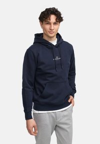 Haut à capuche bleu marine en tissu doux avec une poche kangourou, des poignets et un cordon de serrage. Présente un petit logo sur la poitrine.