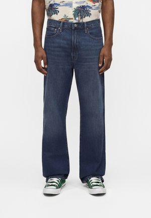 Άνδρας με σκούρα μπλε άνετο τζιν, πράσινα και λευκά αθλητικά παπούτσια και πουκάμισο με τροπικό μοτίβο, στέκεται μπροστά σε απλό λευκό φόντο.