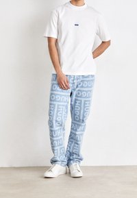T-shirt bianca in cotone con un piccolo logo blu, abbinata a jeans azzurri chiari con un motivo ripetitivo in bianco "HUGO", completata da sneakers bianche.