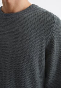 Dark green knitted sweater with a textured pattern, round neckline, and ribbed detailing at the collar and cuffs. Close-up view.