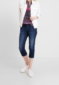 Žena v tmavě modrých zkrácených džínách, bílých teniskách, bílém denimovém kabátu a vzorovaném červeném a námořnickém topu na jednobarevném pozadí.