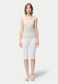 Ung kvinna stående, klädd i ljusgrå ärmlös topp, vita byxor till knäna och beige sandaler, mot en enkel ljus bakgrund.