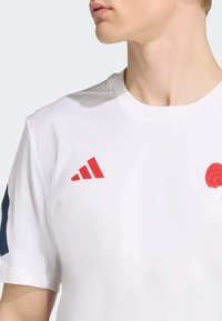 Camiseta deportiva blanca con el logo rojo de Adidas y un emblema circular en el pecho. Diseño de cuello redondo con mangas cortas y tejido texturizado.