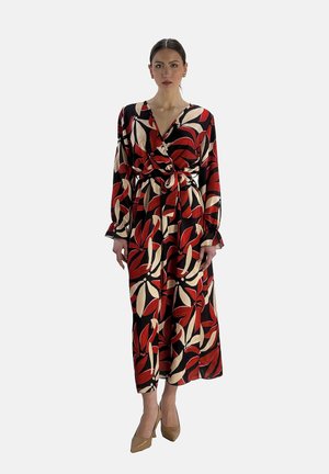 Robe longue à motifs floraux, avec une base noire ornée de motifs rouges et crème audacieux, des manches longues et une taille nouée. Associée à des talons nude.