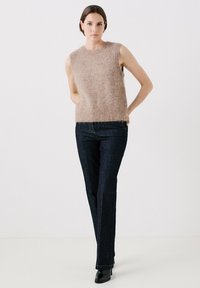 Lichtbruine mouwloze gebreide trui met een zachte textuur, gecombineerd met donkerblauwe rechttoe-rechtaan jeans en zwarte enkellaarsjes.