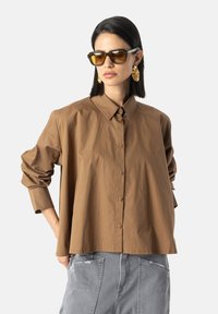 Bruine oversized shirt met knoopsluiting, geplooide mouwen en een relaxte pasvorm, gecombineerd met grijze broek en stijlvolle geelgetinte zonnebril.