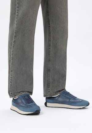 Persona che indossa jeans grigi e sneakers blu con suola bianca, in piedi su uno sfondo bianco con i piedi leggermente divaricati.