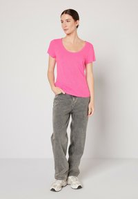 Camiseta rosa de manga corta con escote redondeado, combinada con jeans de cintura alta en gris claro y zapatillas blancas, de pie frente a un fondo neutro.