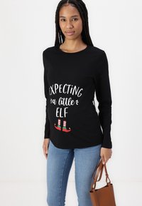 Black long-sleeve shirt with white text saying "Expecting a little Elf," featuring colorful elf shoes at the bottom. Worn with blue jeans and a brown bag.