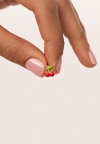 Kirschförmiger Steckohrring mit roten Kirschen und einem grünen Blatt, gehalten von einer Hand mit hellrosa Nagellack vor einem neutralen Hintergrund.