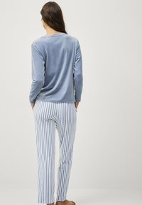 Vrouw in een lichtblauwe lange mouwen fluwelen top en blauw-witte verticaal gestreepte broek, blootvoets staand, met de rug naar ons gekeerd en handen in haar zakken.