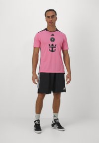 Maglietta atletica rosa con accenti neri e logo, abbinata a pantaloni corti neri con strisce bianche e sneakers nere.