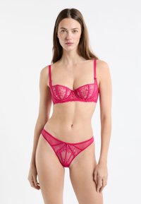 Różowy koronkowy biustonosz i pasujące majtki stringi wykonane z elastycznego materiału, zdobione kwiatowymi wzorami oraz dekoracyjnymi kokardkami na ramiączkach i pasku w talii.