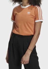 En orange t-shirt med kort ärm och vit ribbad krage, med tre vita ränder på ärmarna och en liten Adidas-logga på bröstet. Kombineras med svarta byxor.