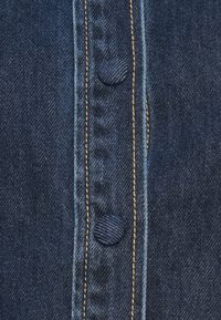 Dark denim fabric with a vertical button placket, featuring a round button and contrasting gold stitching along the seams.