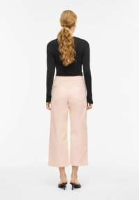 Femme aux longs cheveux bouclés attachés en queue de cheval, portant un haut noir à manches longues, un pantalon large rose clair et des chaussures à talons noires, vue de dos.