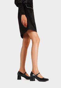 Scarpe con tacco Mary Jane in pelle verniciata nera, con punta tondeggiante, cinturino alla caviglia e tacco a blocco, presentate insieme a un abito nero.