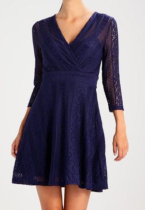 Granatowa koronkowa sukienka z dekoltem w serek, rękawami za łokieć, dopasowaną talią i rozkloszowanym dołem ozdobionym geometrycznymi wzorami.