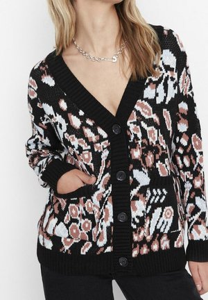 Kobieta ubrana w czarny kardigan zapinany na guziki z białymi i brązowymi abstrakcyjnymi kwiatowymi wzorami, z jedną ręką w kieszeni i srebrnym naszyjnikiem z łańcuszka.