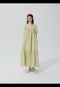 Vrouw die staat in een gele geruite jurk tot op de enkel met opgeblazen mouwen, witte schoenen draagt en een rood haarband.