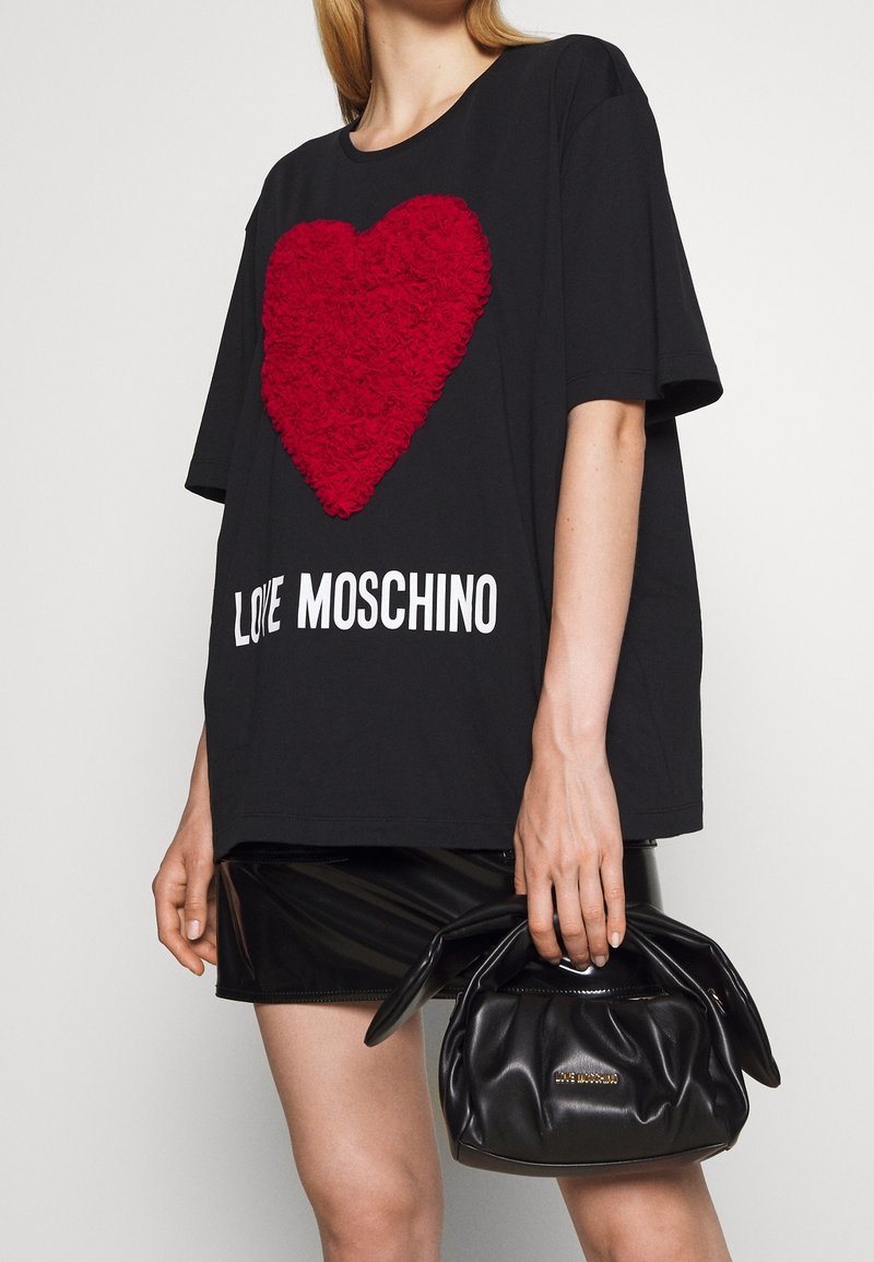 Femme portant un tee-shirt noir oversize avec un cœur texturé rouge et le texte "Love Moschino", tenant un sac à main noir froncé avec des poignées nouées.