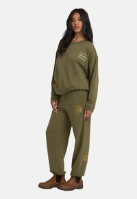 Oliwkowy sweter i dopasowane spodnie wykonane z miękkiego materiału. Sweter posiada logo i kieszenie; spodnie mają wzory słońca blisko dołu.