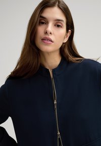 Dunkelblaue Zip-Jacke mit hohem Kragen, aus glattem Stoff, mit einem goldenen Reißverschluss und minimalistischer Gestaltung. Keine zusätzlichen Verzierungen.