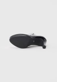 Mocassin à talon pour femme en cuir verni noir vu de la semelle sur fond blanc.
