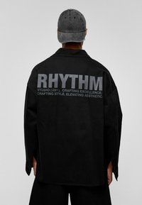 Zwarte oversized shirt met een matte textuur, voorzien van grote grijze "RHYTHM" tekst op de achterkant en subtiele detaillering op de mouwen. De pet is in dezelfde kleur.