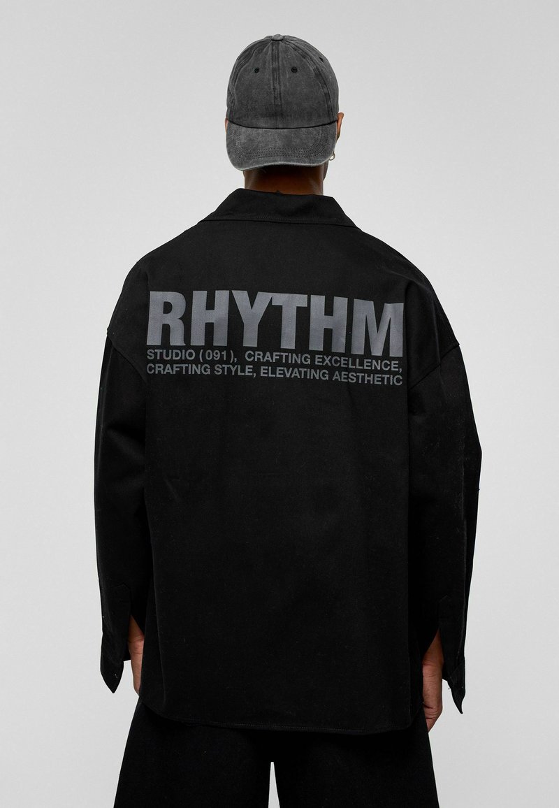 Zwarte oversized shirt met een matte textuur, voorzien van grote grijze "RHYTHM" tekst op de achterkant en subtiele detaillering op de mouwen. De pet is in dezelfde kleur.