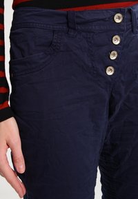 Námořnické modré kalhoty s jemně zmačkanou texturou, vybavené předním uzavíráním na pět knoflíků, bočními kapsami a designem s mírným pasem.