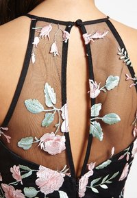 Robe noire avec un dos transparent orné de broderies florales en rose et vert, complété par un détail en forme de trou de serrure et une fermeture à bouton.