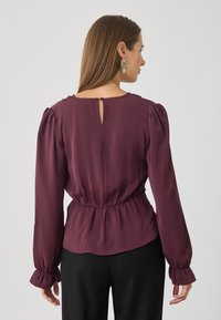 Blusa burdeos con mangas largas y abullonadas, cintura ajustada y detalle de escote en la espalda. Textura suave, tejido ligero. Combinada con pantalones negros.